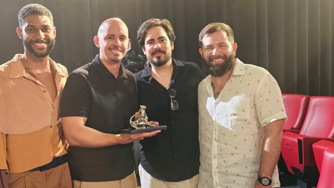 Quatro homens sorrindo, um deles segurando um trof&eacute;u dourado sobre uma base preta.