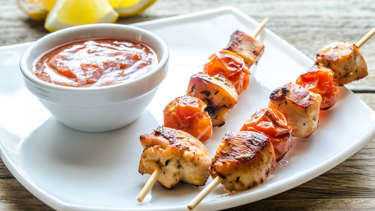Espetinhos de frango e tomate são ótimas opções de petiscos práticos para saborear com as mãos enquanto acompanha os jogos do Brasil na Copa do Mundo. Dois espetinhos de frango e tomate-cereja grelhados, acompanhados de molho em tigela branca.