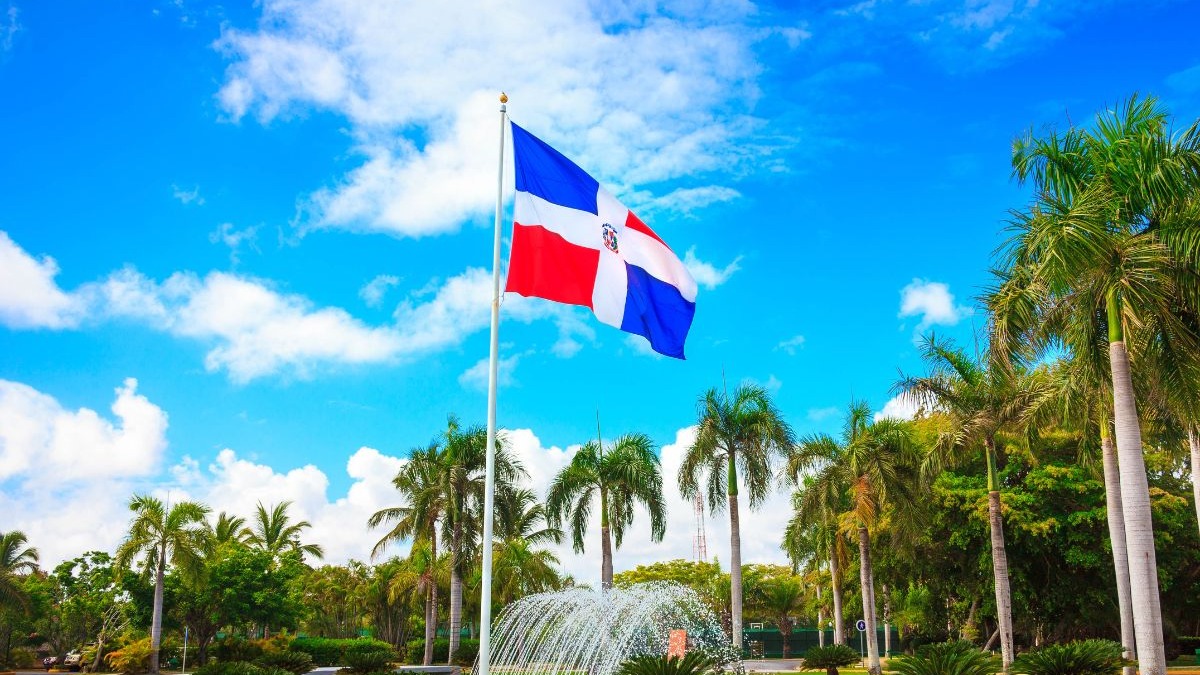 Bandeira da Rep&uacute;blica Dominicana em mastro alto, c&eacute;u azul, nuvens, palmeiras e fonte.