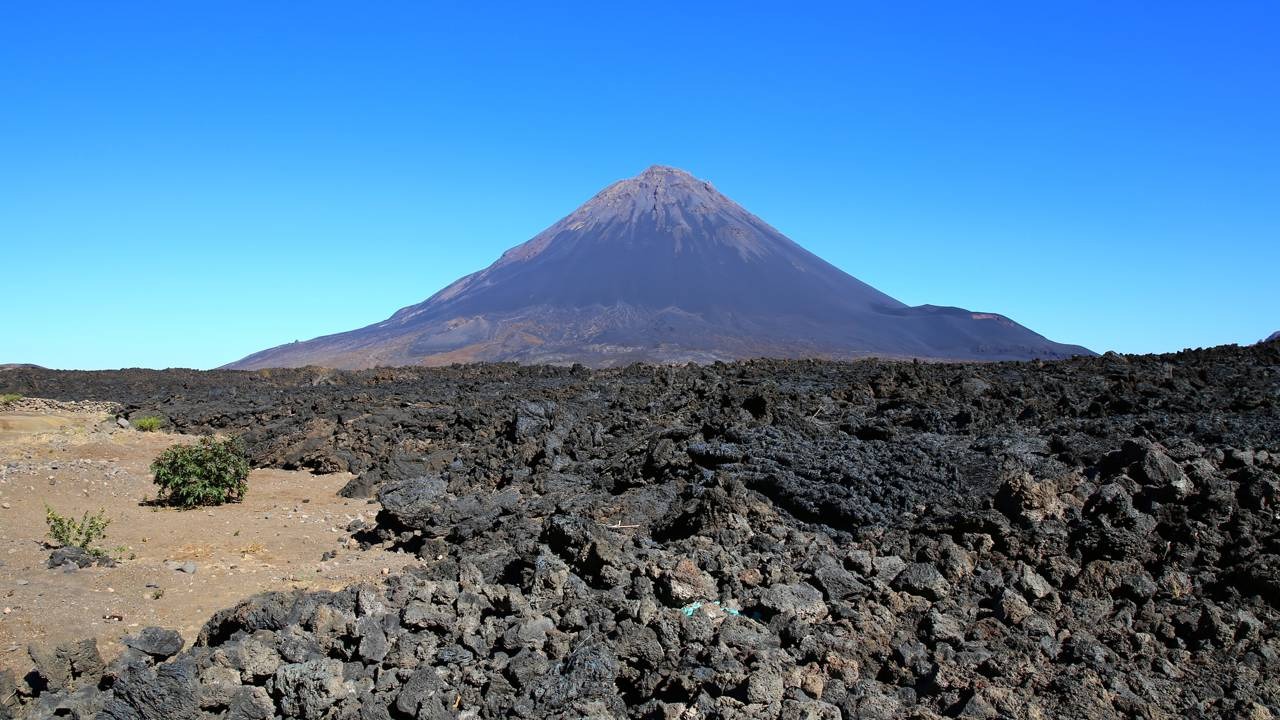 O Pico do Fogo, vulcão ativo de Cabo Verde, é peça central da geografia acidentada do arquipélago destacada na matéria. Pico do Fogo, vulcão cinza e negro, campos de lava vulcânica, céu azul. Vegetação esparsa.