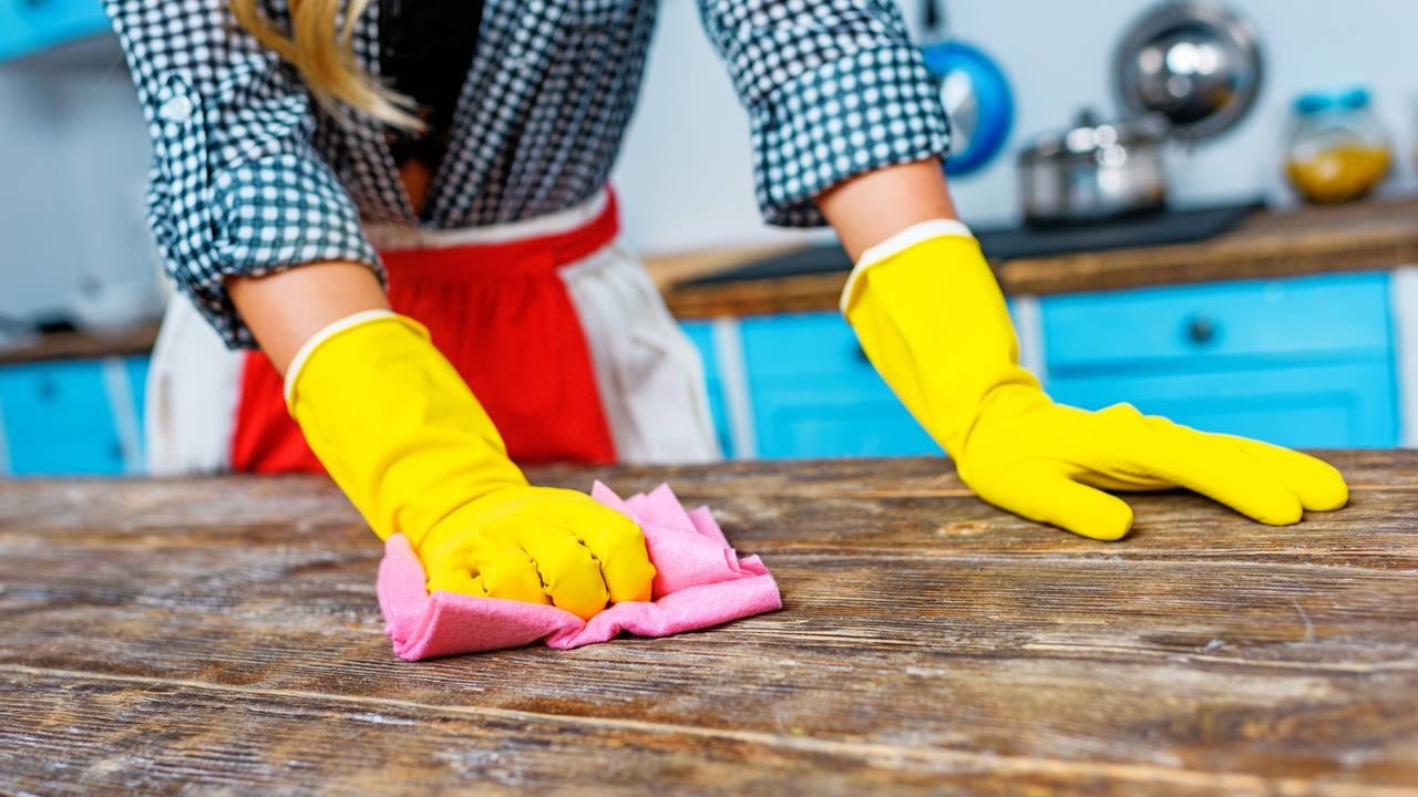 A limpeza regular da cozinha complementa o uso de soluções caseiras para eliminar odores persistentes no ambiente. Pessoa com luvas amarelas limpa uma bancada de madeira com um pano rosa em uma cozinha