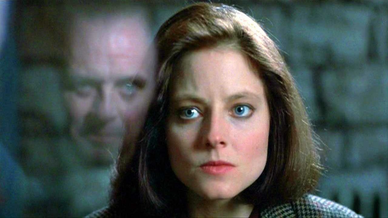 Jodie Foster e Anthony Hopkins em cena icônica de "O Silêncio dos Inocentes", um dos clássicos para curtir no frio. Clarice Starling, à frente, com Hannibal Lecter desfocado ao fundo. Olhar intenso.