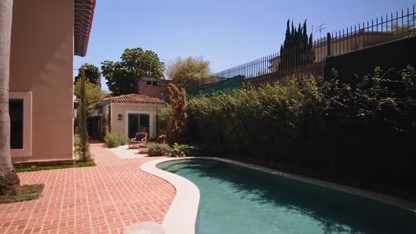 Piscina azul curva, pátio de tijolos, casas, árvores e arbustos sob céu claro