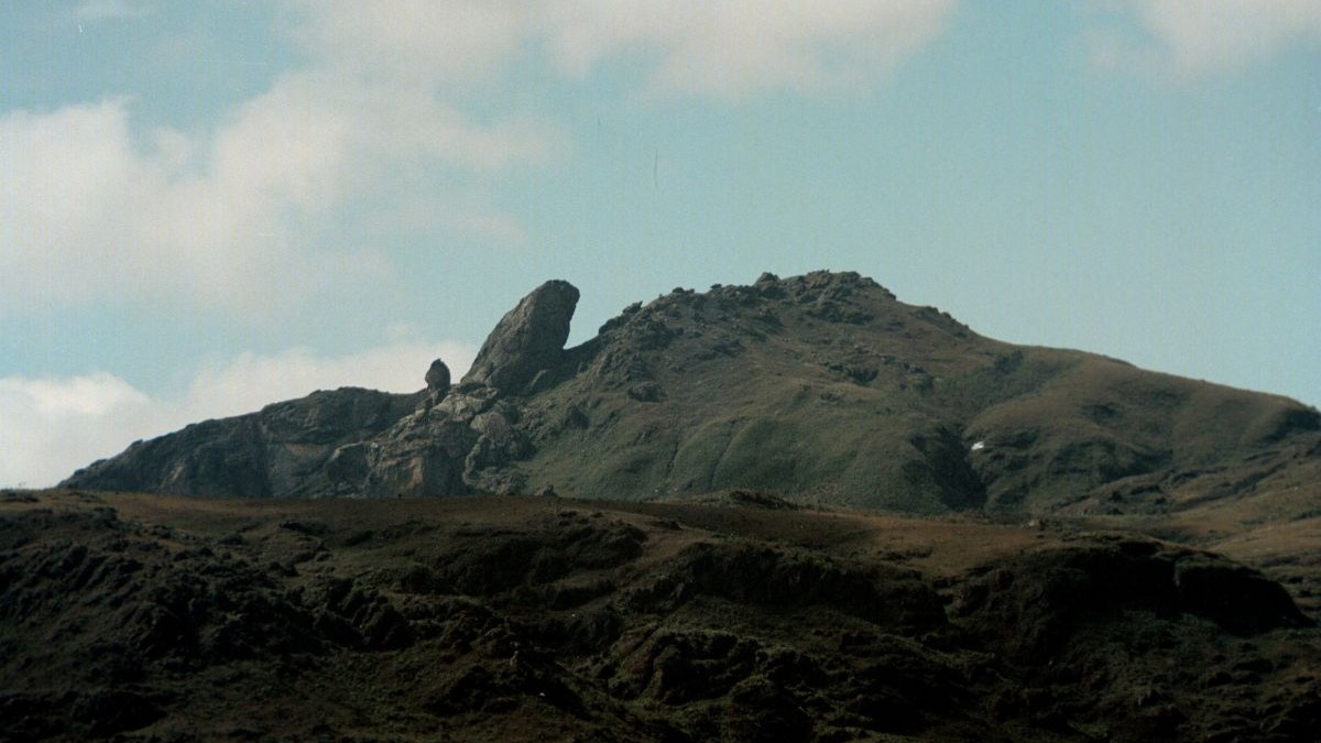 Montanha rochosa com cume e forma&ccedil;&atilde;o de pedra saliente sob c&eacute;u azul com nuvens.