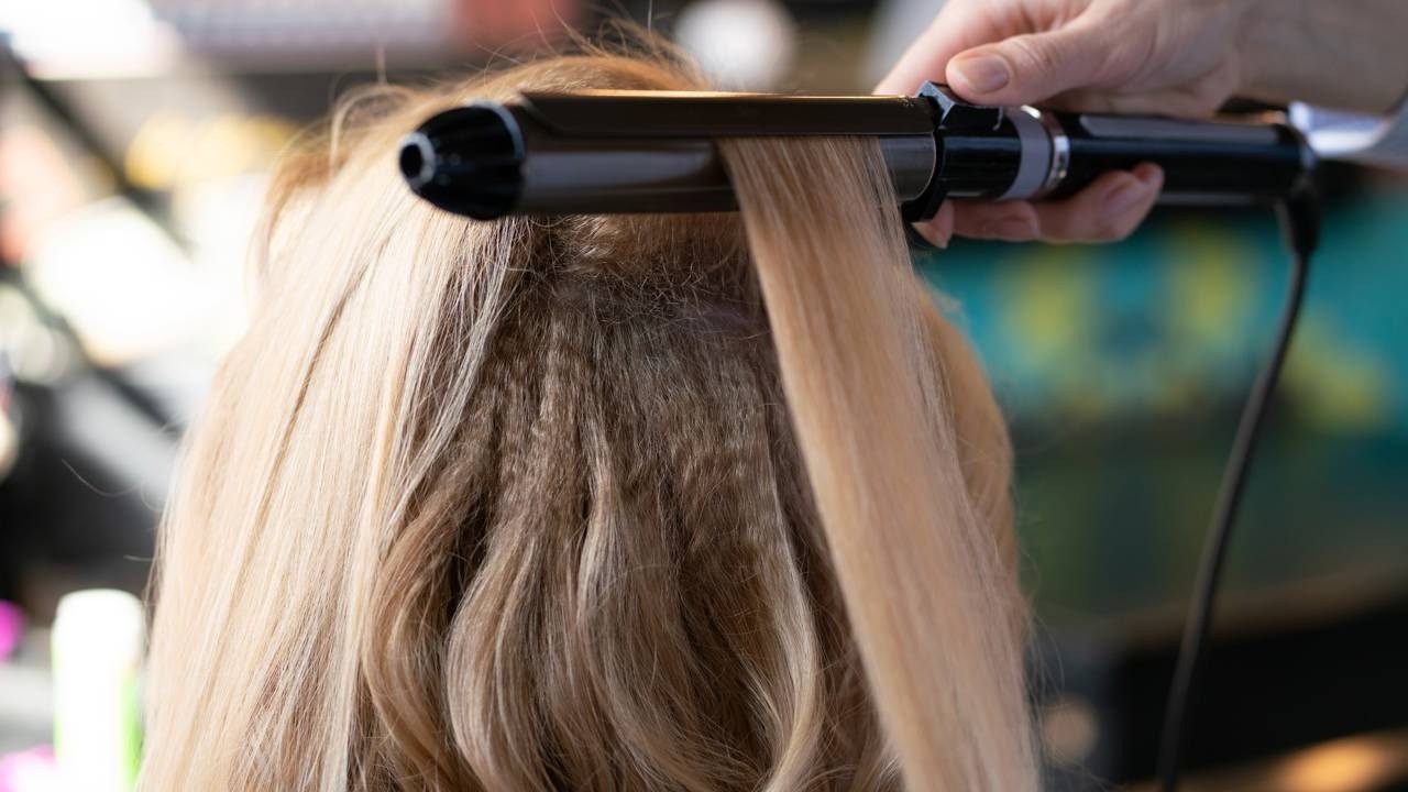 Ferramentas térmicas como modeladores e secadores podem afetar a estrutura do fio e intensificar o frizz, exigindo cuidados extras Mão segura modelador térmico em mecha de cabelo loiro sobre cabelo ondulado