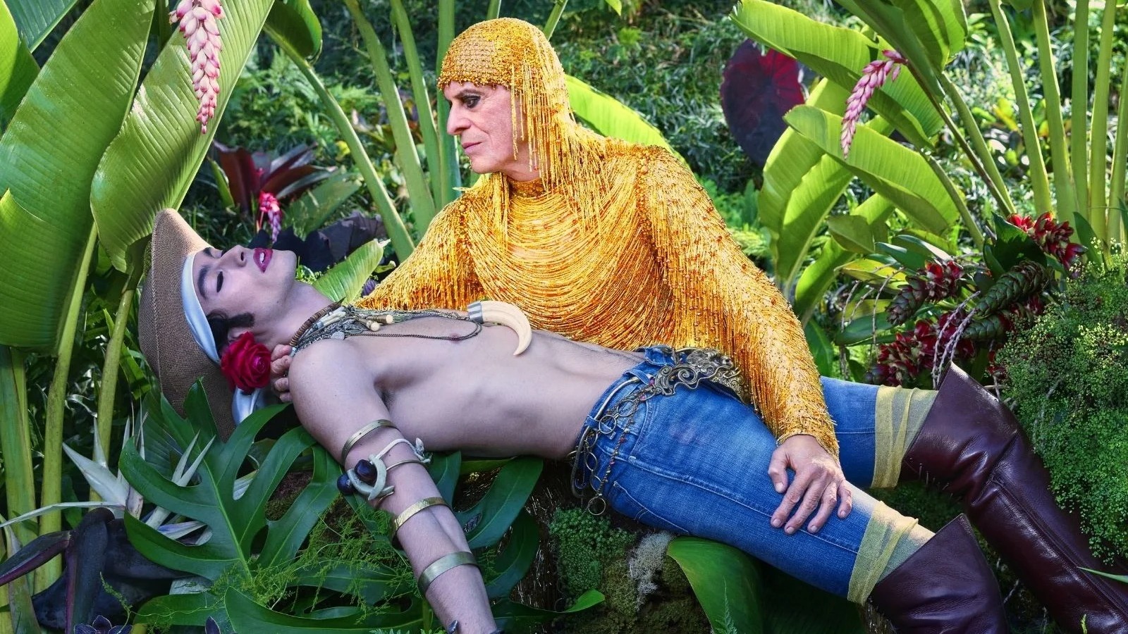 Homem de dourado sobre outro sem camisa com jeans e adere&ccedil;os em jardim tropical