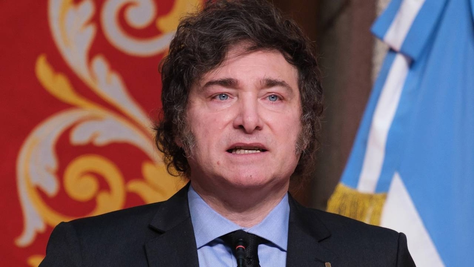 Javier Milei, presidente argentino, defende ajuste fiscal enquanto mudanças na Previdência Social geram insatisfação popular. Javier Milei, de terno e camisa, fala em microfone, com fundo vermelho e bandeira argentina