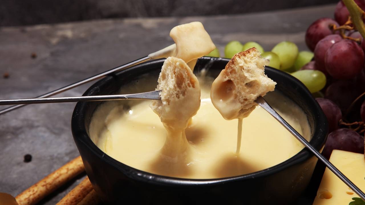 Pães mergulhados no fondue de queijo cremoso, uma das sugestões de acompanhamento para a receita ideal nos dias frios. Garfos com pão e um pedaço de alimento mergulhados em fondue de queijo cremoso