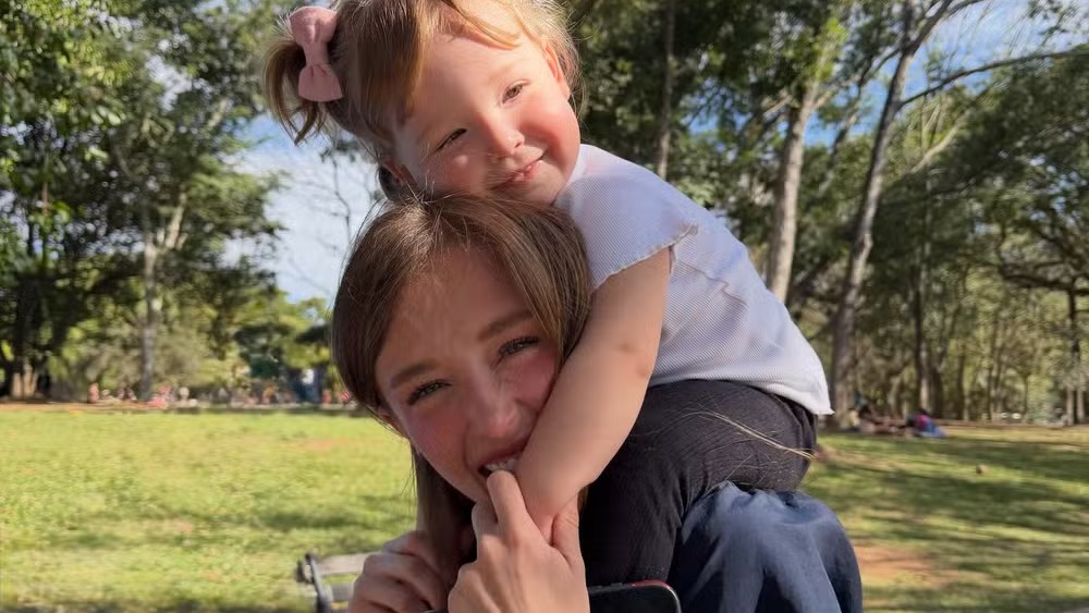 A atriz Duda Reis reforça a importância de atividades ao ar livre e da socialização para a saúde mental. Mulher e criança sorridentes, criança nos ombros da mulher, em parque ensolarado com árvores