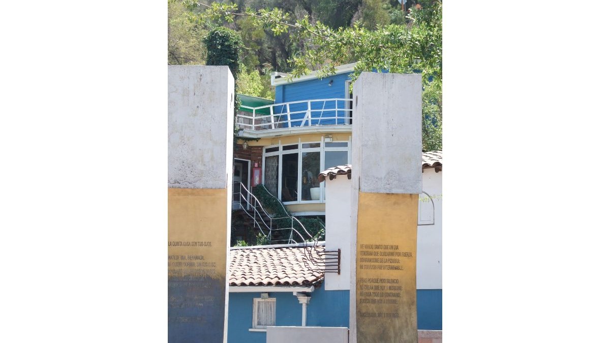 Paredes com poemas emolduram uma casa colorida de vários andares, com janelas e varanda, cercada por árvores.