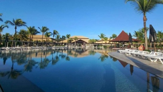 Ampla piscina de resort refletindo céu azul, palmeiras, espreguiçadeiras e edifícios ao fundo.
