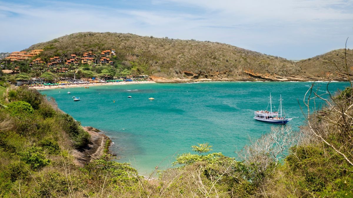 Vista aérea de baía com praia movimentada, mar azul-turquesa, barcos, casas na colina e vegetação.
