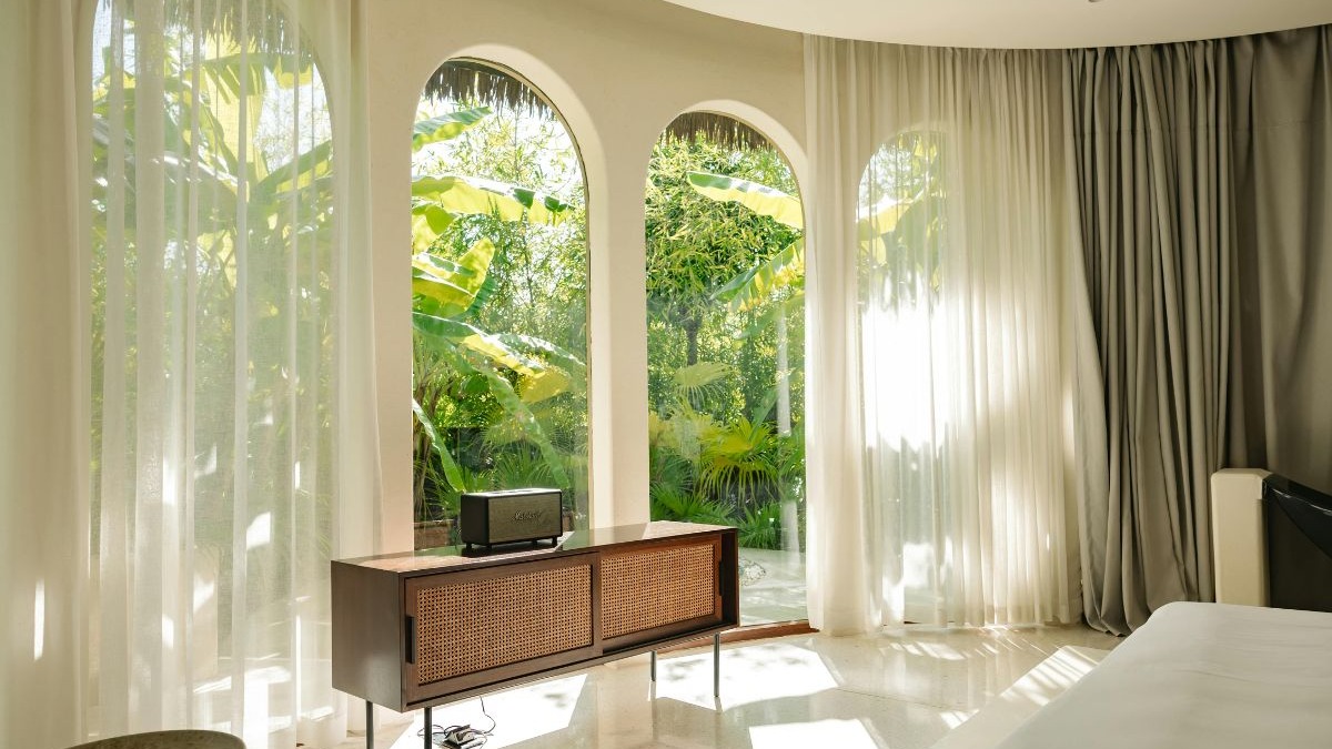 Ambiente com janelas arqueadas, cortinas brancas transl&uacute;cidas, m&oacute;vel de madeira e folhagens verdes.