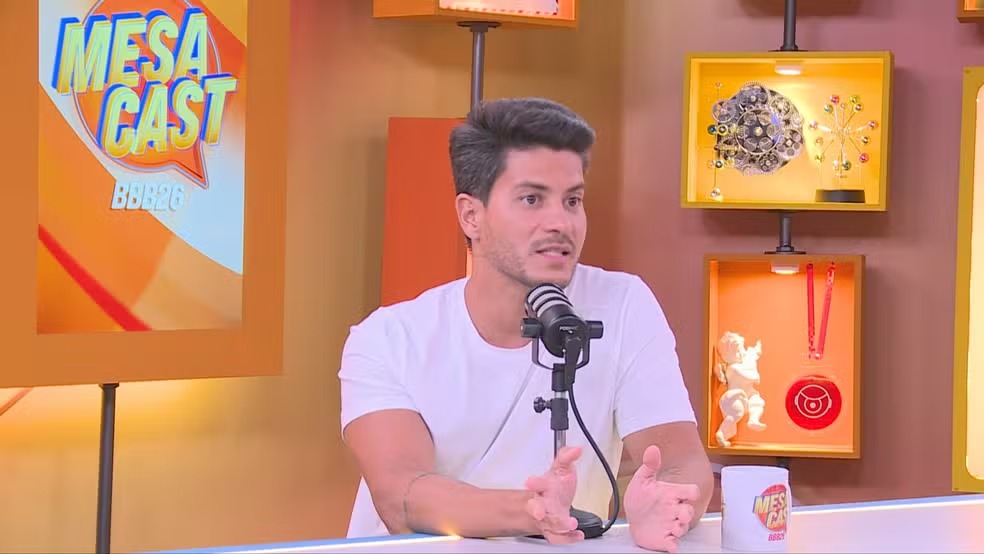 Arthur Aguiar, cuja liderança no BBB 22 foi decisiva para sua vitória, em participação no programa Mesa Cast. Arthur Aguiar, de camiseta branca, fala em microfone no estúdio do Mesa Cast BBB26.