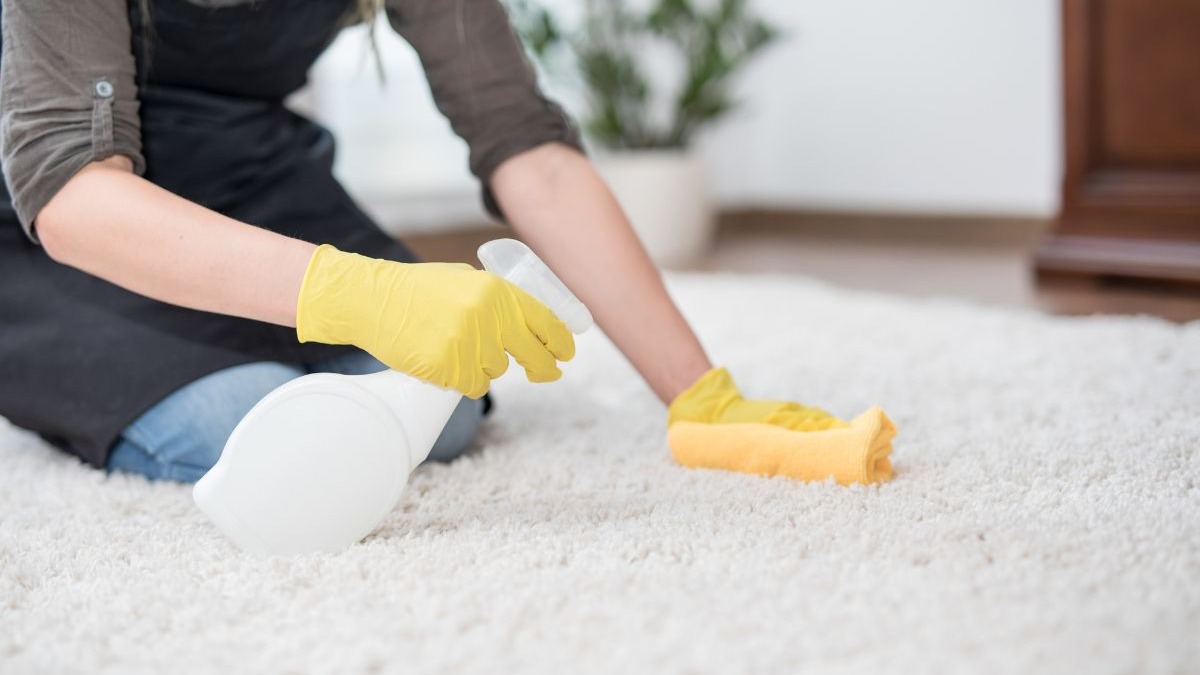 Pessoa de luvas amarelas e avental usando spray e pano para limpar carpete branco.