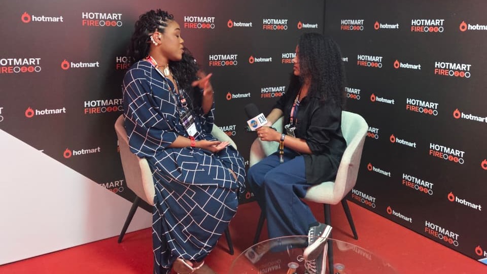 Duas mulheres se entrevistam no Hotmart Fire, uma com microfone, fundo com logos do evento