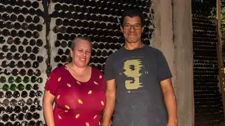Dóris Dias e Robson Lopes mostram o resultado do projeto da casa construída com cerca de 20 mil garrafas de vidro recicladas em Foz do Iguaçu. Casal sorridente em frente a uma parede de casa construída com garrafas de vidro