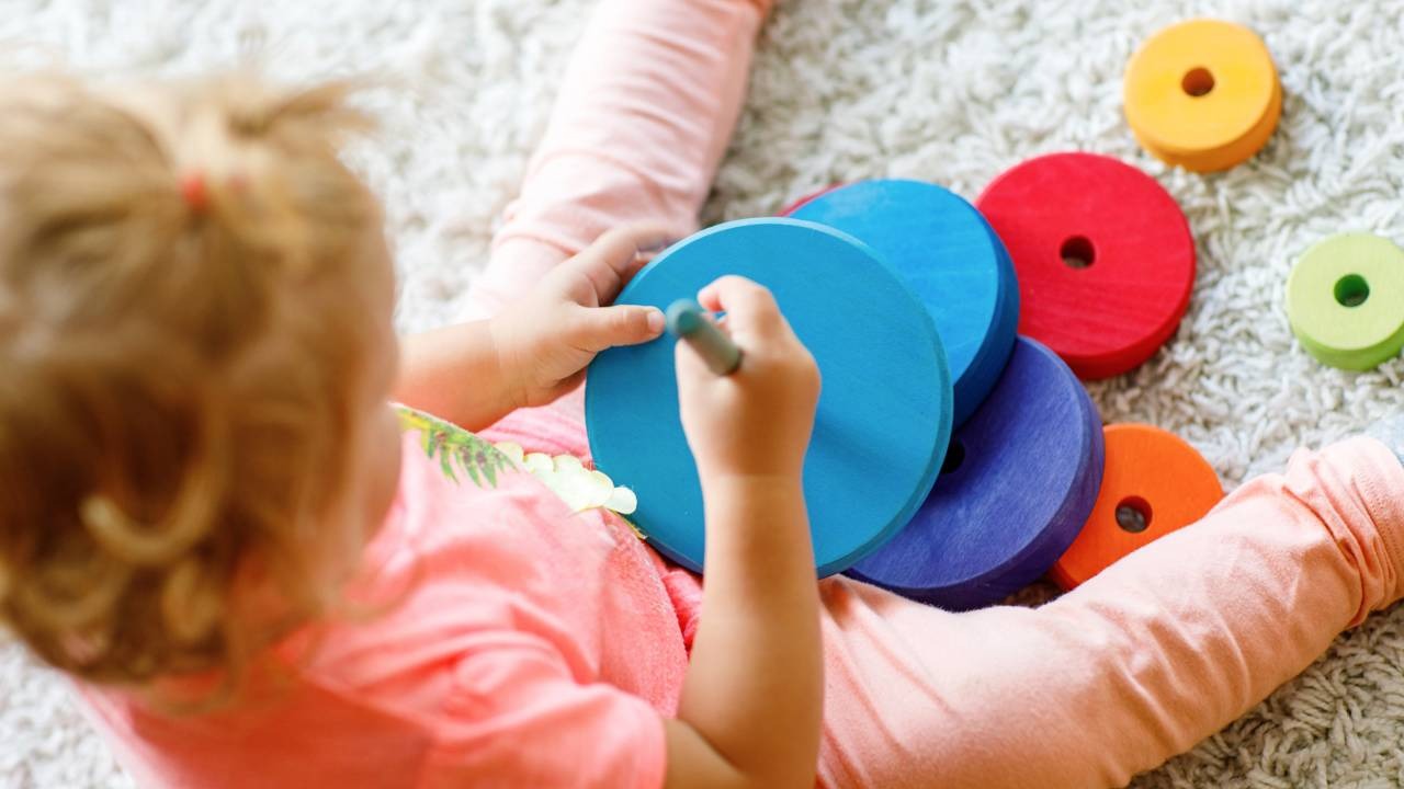 A brincadeira com brinquedos vira uma missão divertida para incentivar a organização das crianças em casa. Mãos de uma criança brincando com argolas coloridas de madeira sobre um tapete.