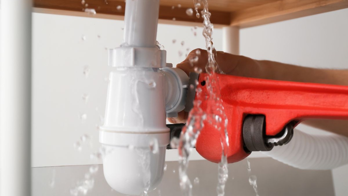 M&atilde;o segurando chave de grifo vermelha em cano de PVC branco vazando &aacute;gua.