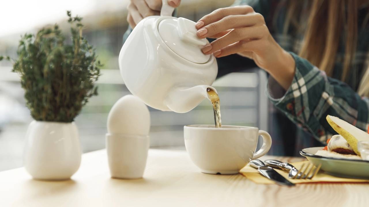 O consumo de chá quente é um dos hábitos que auxiliam no alívio de sintomas de gripes e resfriados. Mãos femininas servem chá quente de bule branco em xícara na mesa com comida e planta