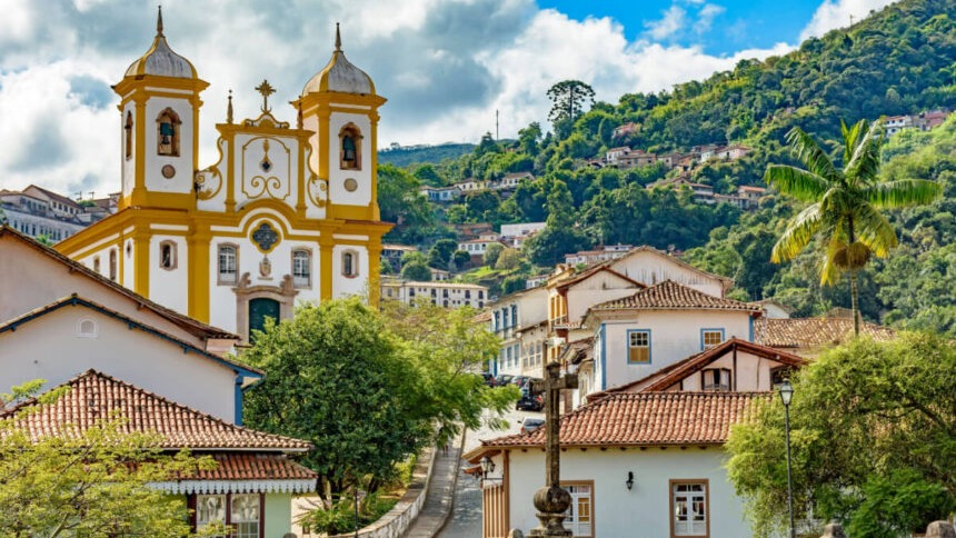 Vista panorâmica de igreja barroca amarela e branca, casas coloniais e morro arborizado.