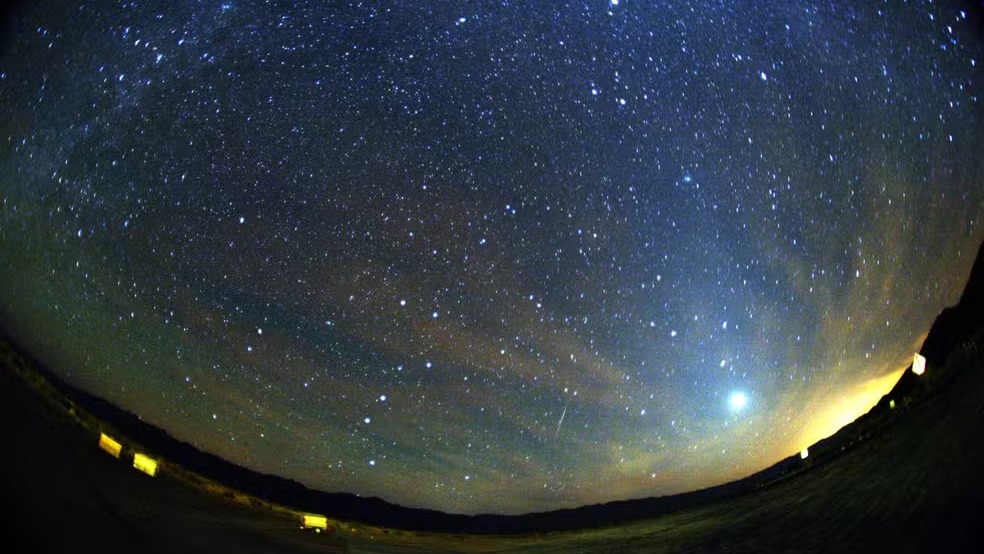 Meteoro da chuva Orionídeas em 2007. Céu noturno estrelado com rastro de meteoro sobre um horizonte escuro e luzes distantes