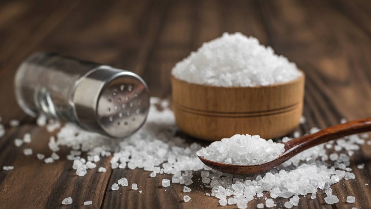 Uma pitada estratégica de sal realça sabores e equilibra doces e cafés, conforme truques culinários revelados. Saleiro, tigela e colher de pau com sal grosso em uma mesa de madeira