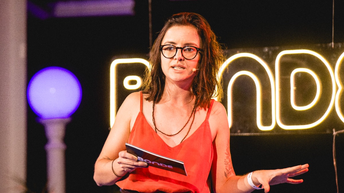 Talita Novacoski em um palco, segurando folheto BONDE, com letreiro neon BONDE ao fundo