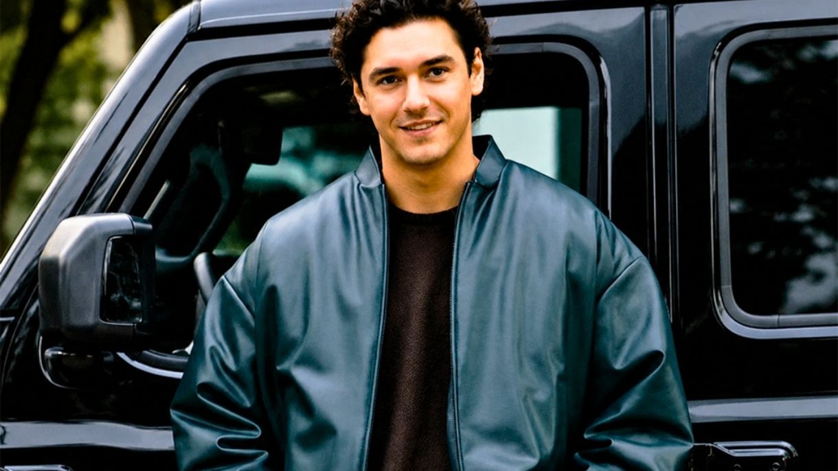 Garrett Graham, da série "Off Campus", exemplifica o estilo casual e esportivo de "golden boy" com sua jaqueta bomber. Homem sorridente, cabelo cacheado, jaqueta bomber azul, suéter marrom, posando junto a carro.