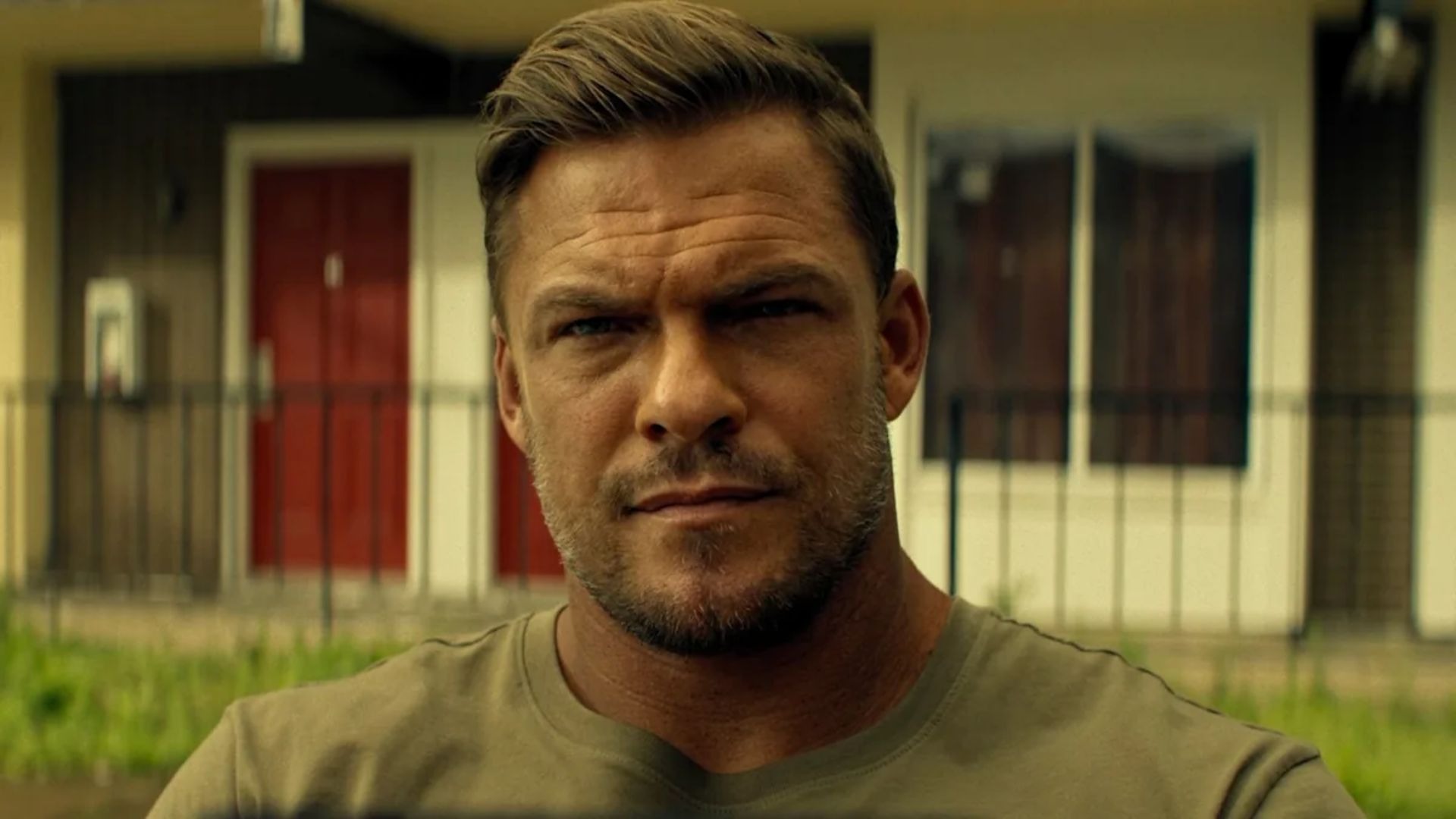 Alan Ritchson, astro de Reacher, em sua jornada para se consolidar como um dos principais nomes do cinema de ação. Alan Ritchson olhando sério, cabelo e barba curtos, camiseta verde, casa desfocada ao fundo