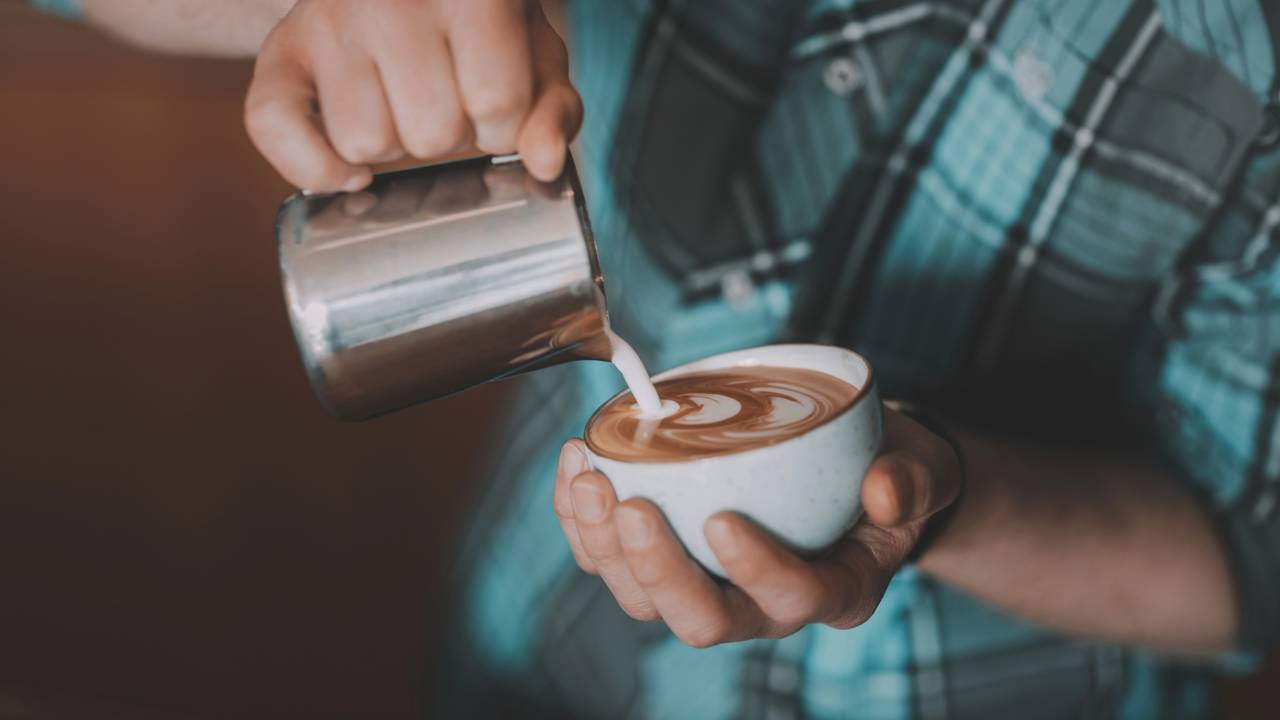 Aprenda a preparar seu próprio cappuccino caseiro com sabor e cremosidade de cafeteria para desfrutar a qualquer momento. Mãos despejando leite vaporizado de jarra de metal em xícara de café com arte latte