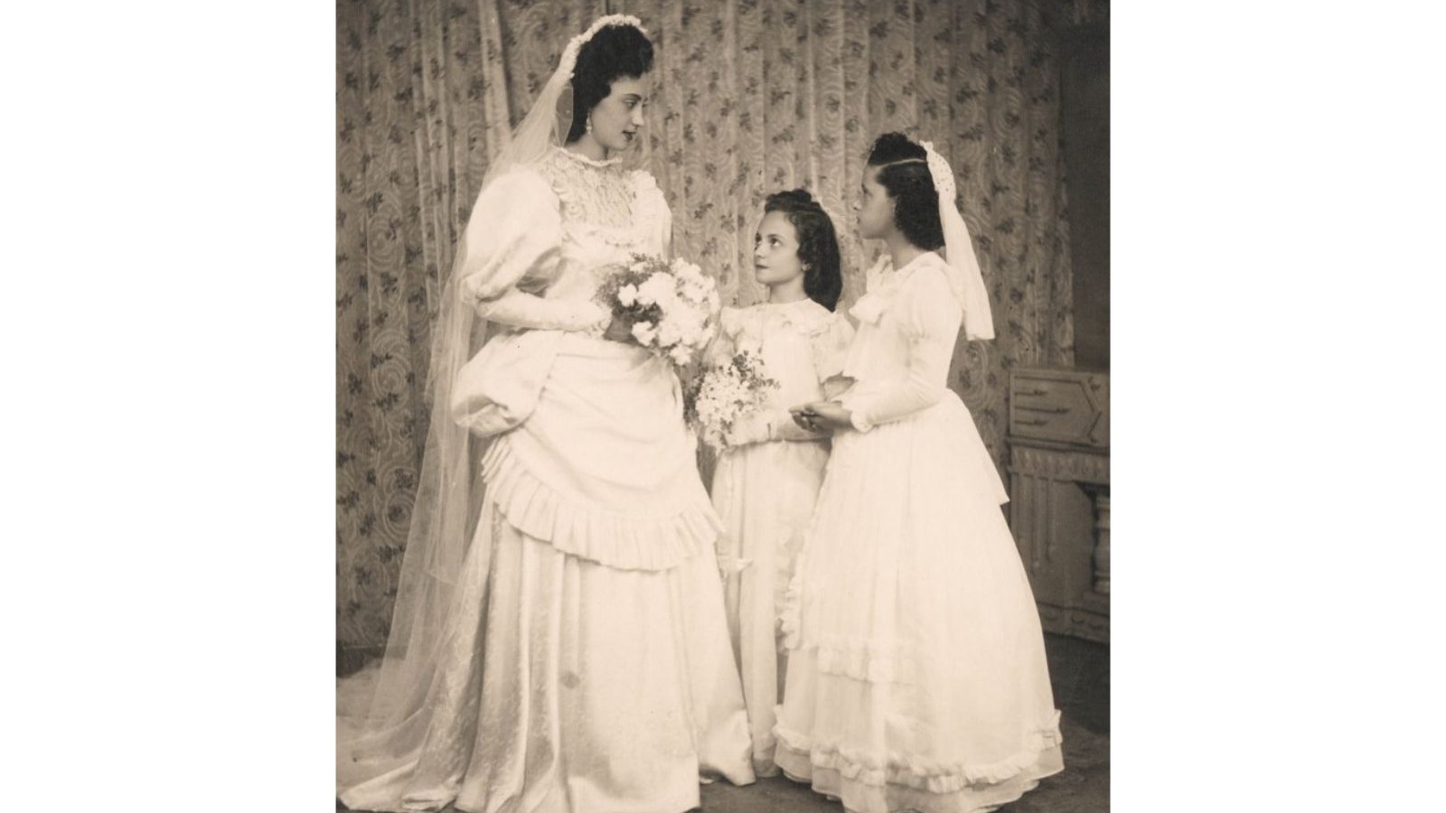 Tr&ecirc;s mulheres em vestidos de casamento vintage, uma adulta e duas crian&ccedil;as segurando buqu&ecirc;s