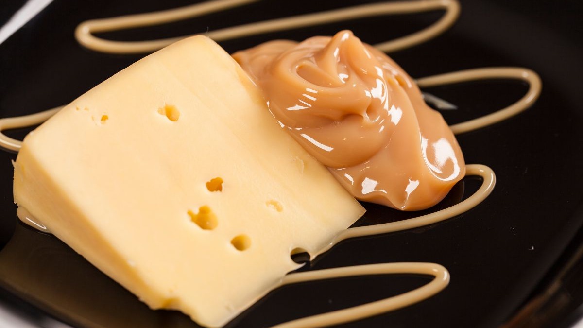 Doce de leite com queijo Minas: a combinação clássica mineira para fechar as refeições e aquecer o inverno. Fatia de queijo com furos e doce de leite cremoso em prato escuro.