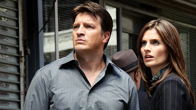 Nathan Fillion e Stana Katic em cena. Ele de camisa listrada, ela de blazer pinstripe.