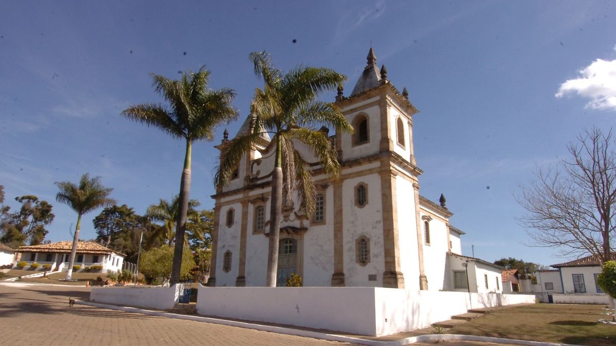 Igreja colonial branca com torres, palmeiras e c&eacute;u azul, em charmoso vilarejo mineiro.