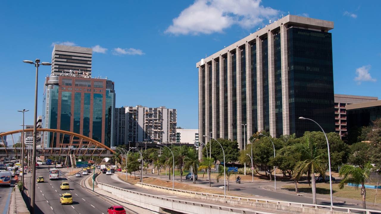 A Prefeitura do Rio é responsável por serviços como escolas municipais, unidades de saúde e transporte público municipal. Vista panorâmica de cidade com edifícios altos, avenida movimentada, ponte, árvores e céu azul.