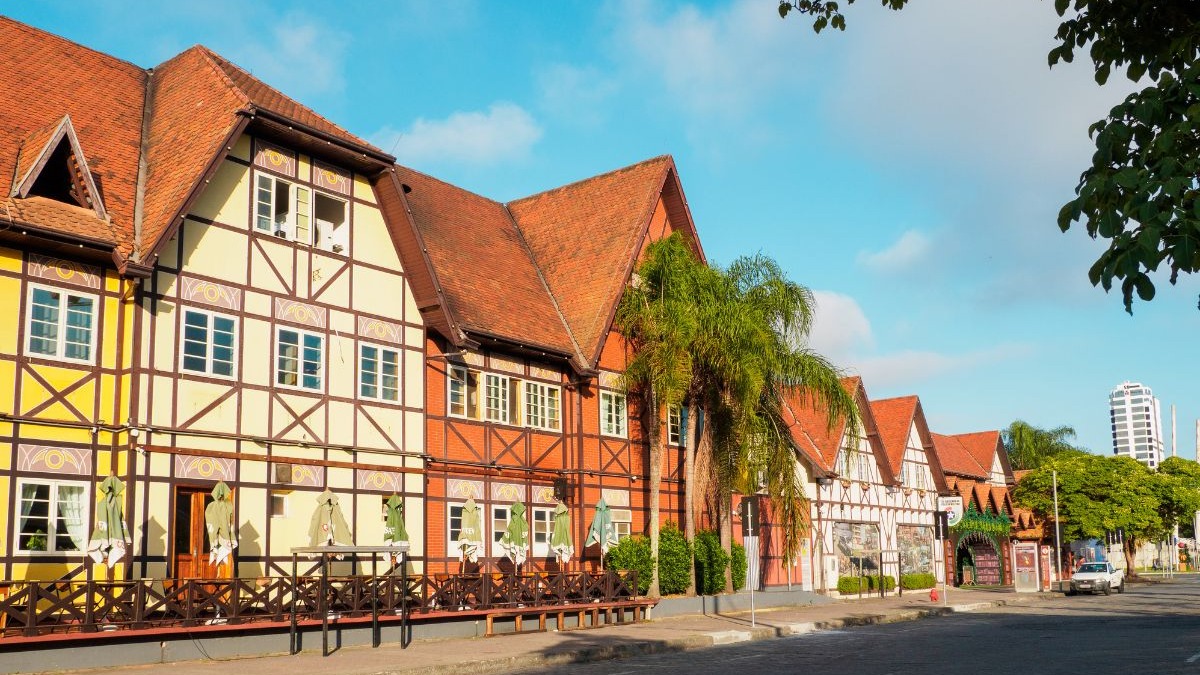 Fileira de casas com arquitetura enxaimel de telhados vermelhos sob c&eacute;u azul, com palmeiras.