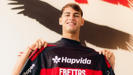 Joaquín Freitas posa com a camisa do Flamengo, reforço argentino anunciado oficialmente pelo clube. Foto: Adriano Fontes/CRF Homem jovem sorridente segura camisa preta e vermelha Hapvida FREITTAS contra parede clara