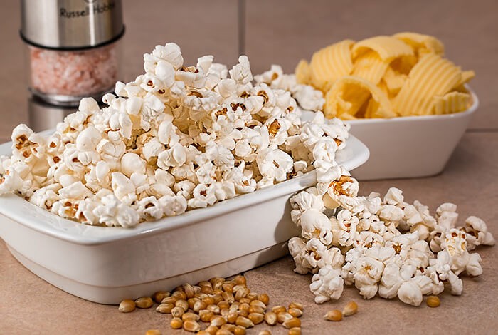 A bowl of popcorn, a bowl of crackers, a Himalayan salt grinder, and corn kernels.