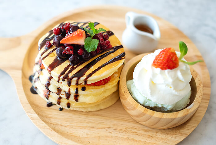 A stack of pancakes with fruit toppings, choco syrup, whipped cream, and maple syrup on the side.