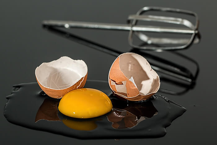 A cracked egg on a surface with an egg beater in the background.