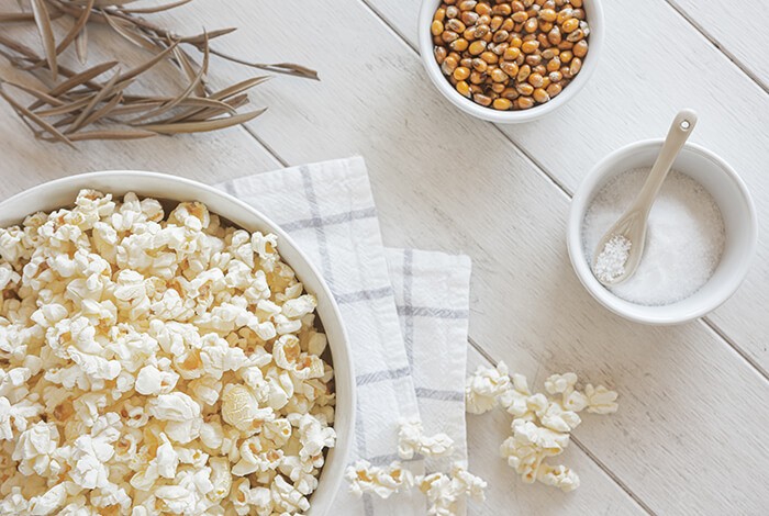 A bowl of popcorn, a bowl of salt, a bowl of corn kernels, a hand towel, and dried leaves on top of a white wooden surface.