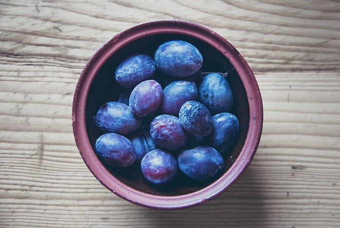 Plums placed in a clay bowl on top of a wooden surface. 