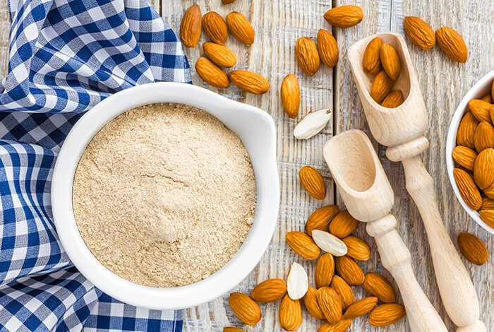 Almond flour in a bowl and a handful of almonds scattered on a wooden surface.