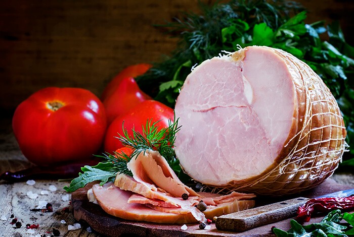 Sliced ham and knife on top of a wooden chopping board with tomatoes in the background.