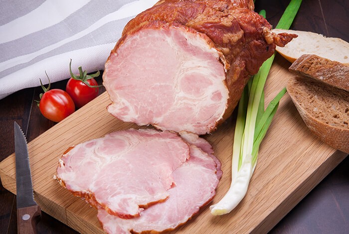 Sliced ham, sliced bread, a bread knife, tomatoes, and green onions on top of a wooden chopping board.