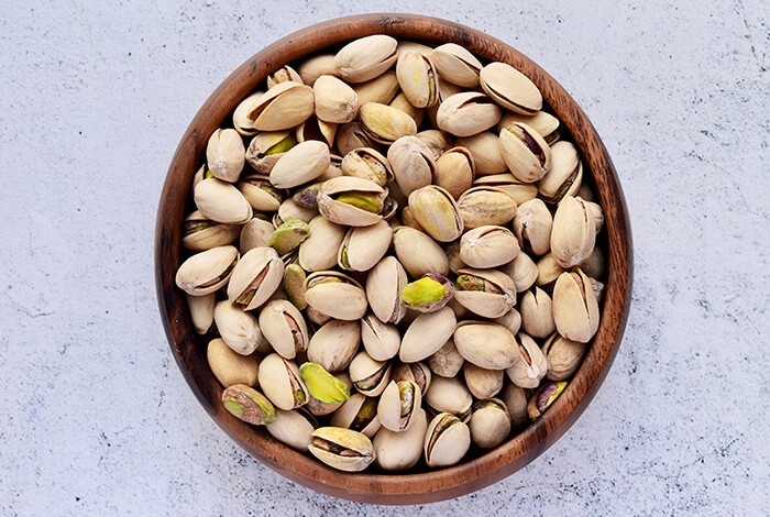 A wooden bowl of pistachios on a rough surface.
