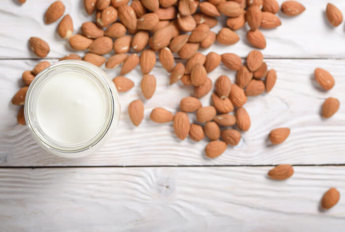 A glass of almond milk and a handful of almonds scattered on a white wooden surface.