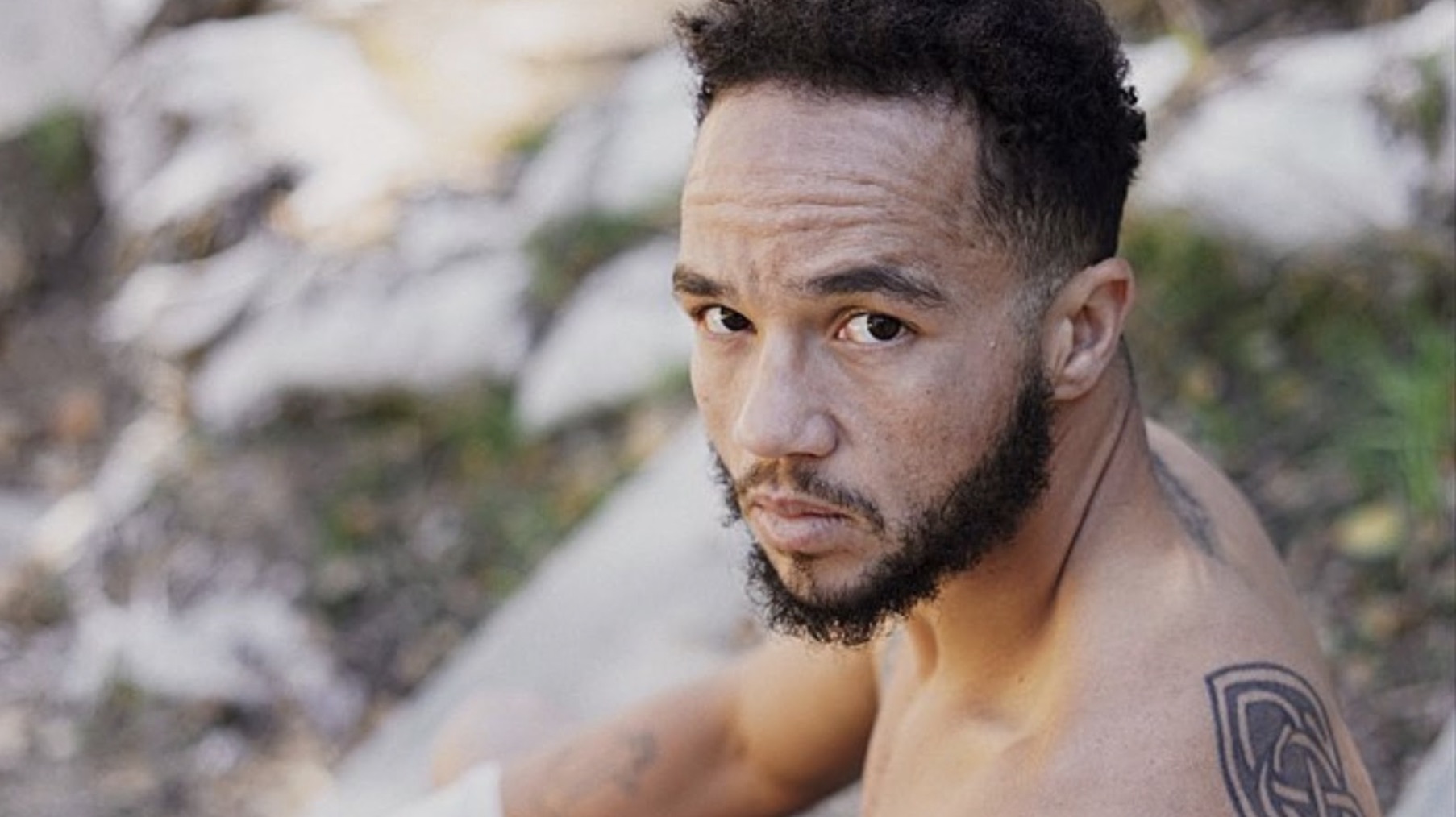 Patricio Manuel is posing for a photo. He is sitting down and facing the camera. His top is off, and he has tattoos on his back and arms.