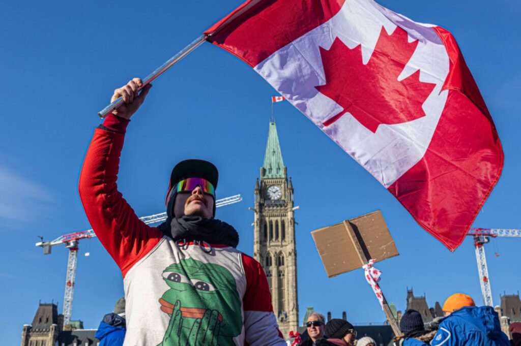 A man dressed in winter gear holds a Canadian flag in the air during a protest